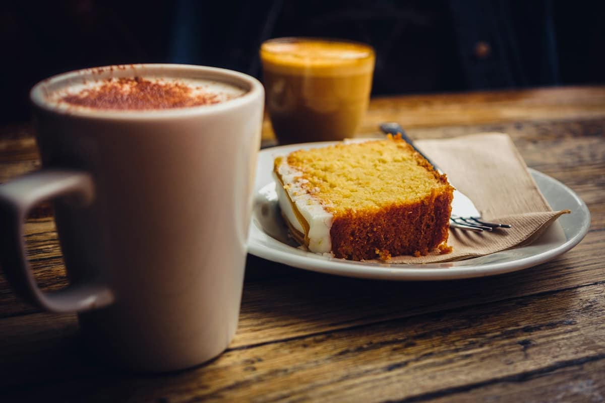 A slice of cake on a plate beside a mug, on a wooden table