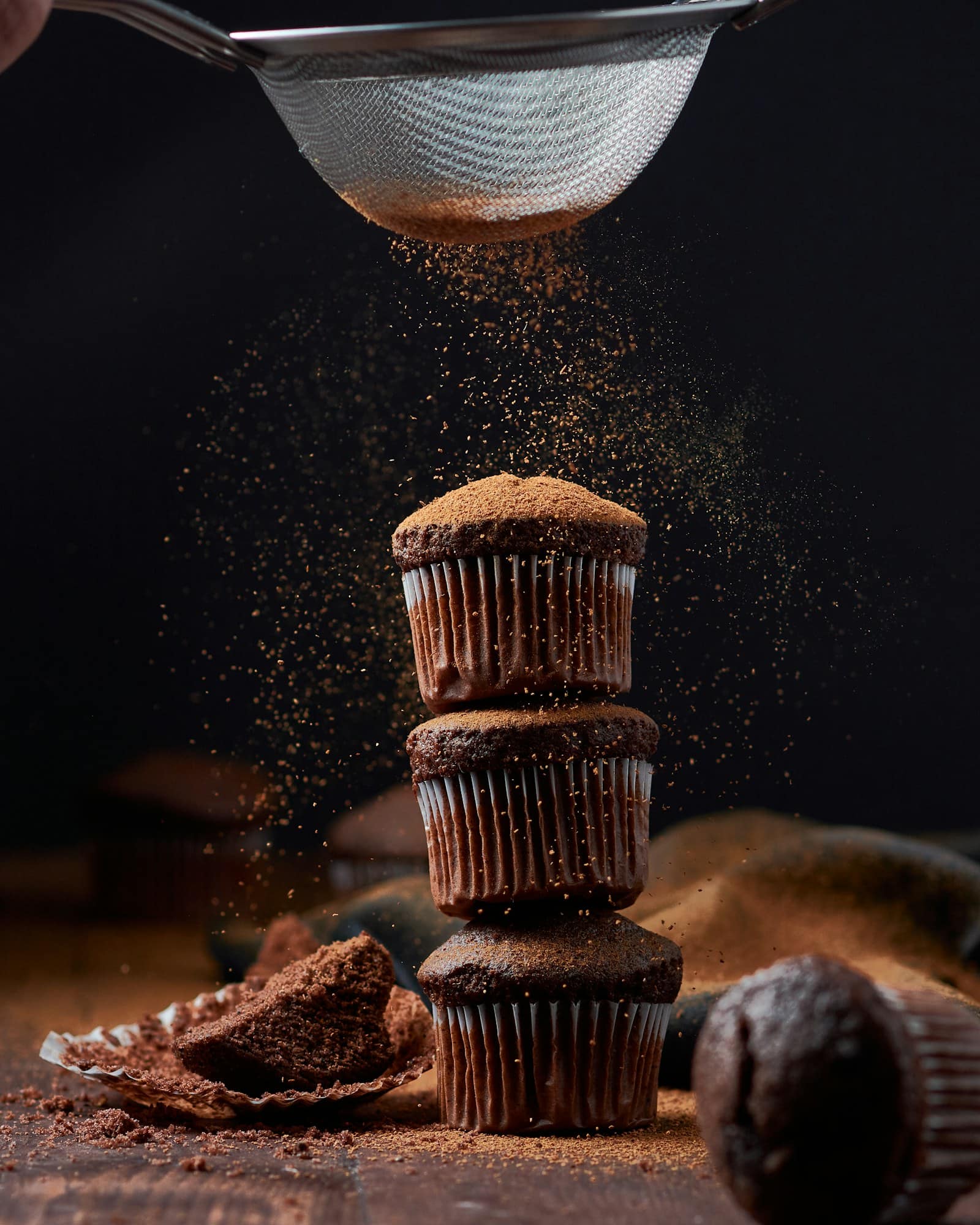 A stack of chocolate muffins being dusted with cocoa through a sieve, lit low against a dark background
