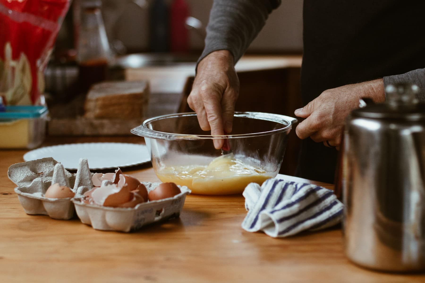 A home baker whisking eggs in a glass bowl on a wooden kitchen counter